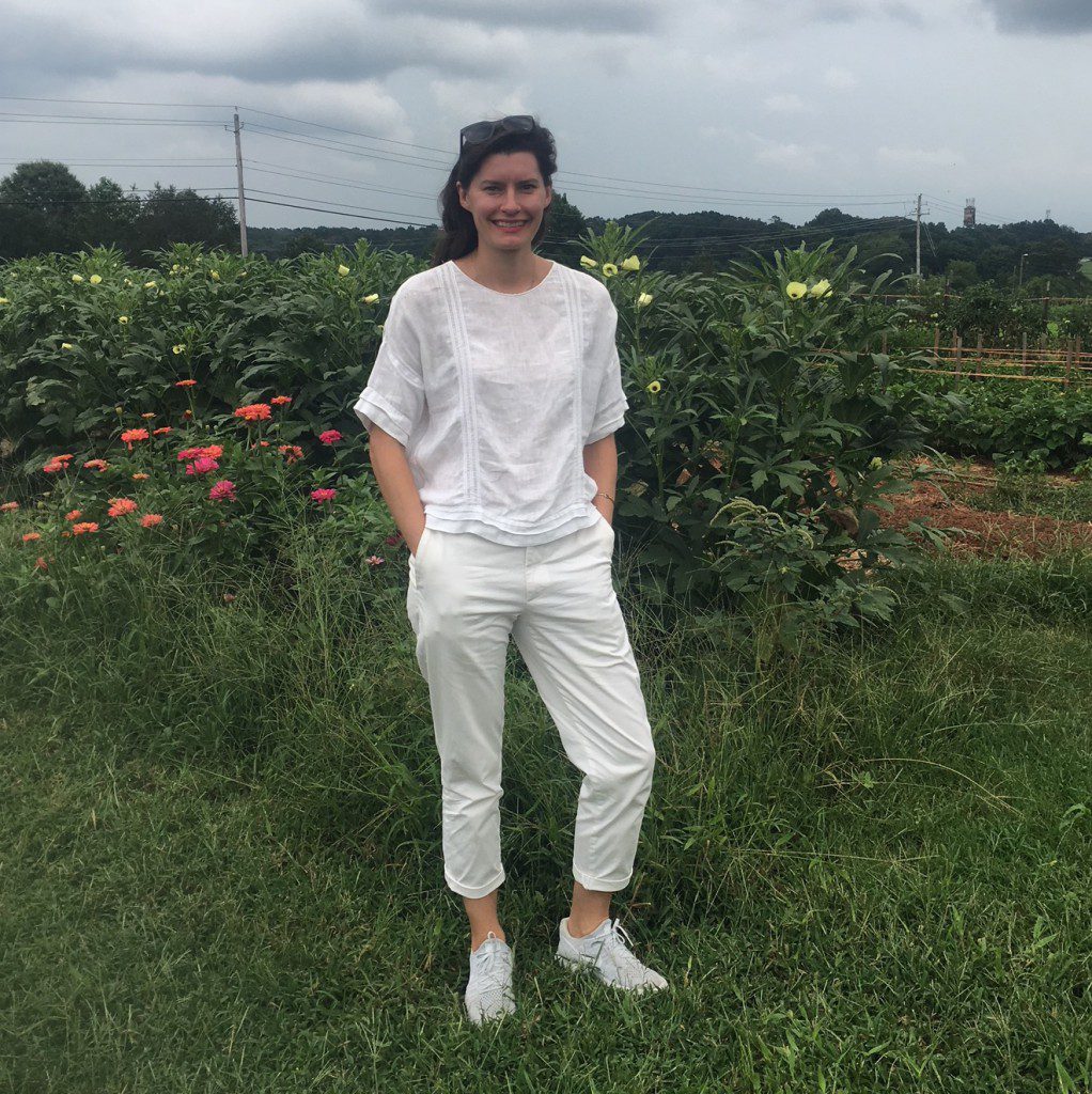 A woman wearing all white stands on a farm in front of bushy green plants.