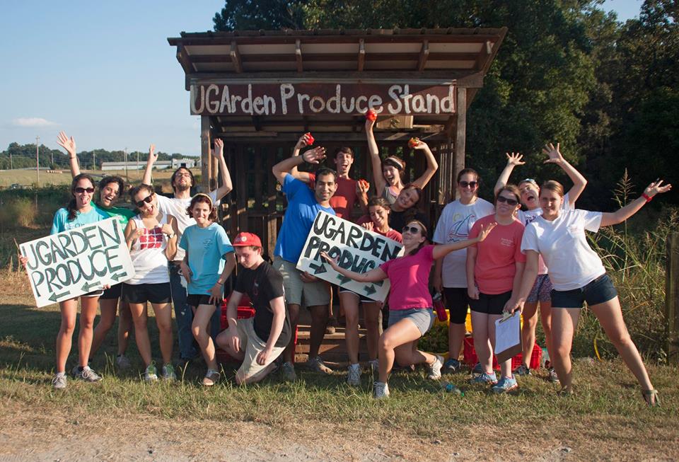 A group of people stand outside of a small building with a sign that says "UGArden Produce Stand."